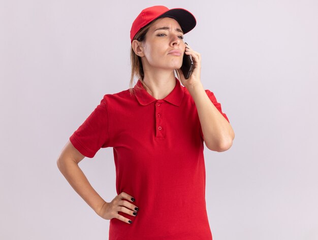 Déçu jeune jolie femme de livraison en uniforme parle au téléphone et regarde à côté isolé sur mur blanc