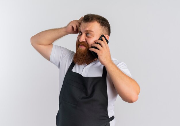 Déçu professionnel barbu homme barbu en tablier de parler au téléphone mobile à côté de la tête de grattage confus debout sur un mur blanc