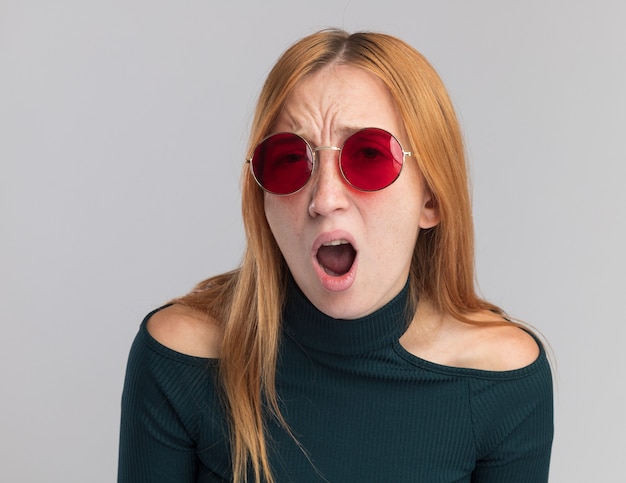 Déçue jeune fille rousse au gingembre avec des taches de rousseur dans des lunettes de soleil isolées sur un mur blanc avec espace de copie