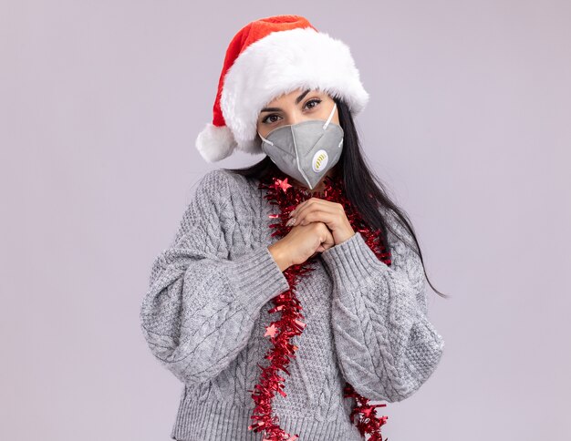 délicate jeune fille caucasienne portant un chapeau de noël et une guirlande de guirlandes autour du cou avec un masque de protection gardant les mains ensemble isolées sur un mur blanc avec espace de copie