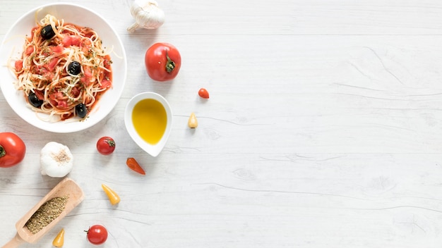 De délicieuses pâtes à spaghetti sur une assiette; tomate fraîche; bol d&#39;huile d&#39;olive et d&#39;herbes sur un bureau en bois