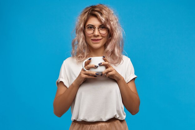 Délicieux. Portrait de jolie jeune femme ringard portant des lunettes rondes bénéficiant d'un chocolat chaud sucré. Jolie fille aux cheveux rosés tenant une tasse blanche, buvant du bon café frais