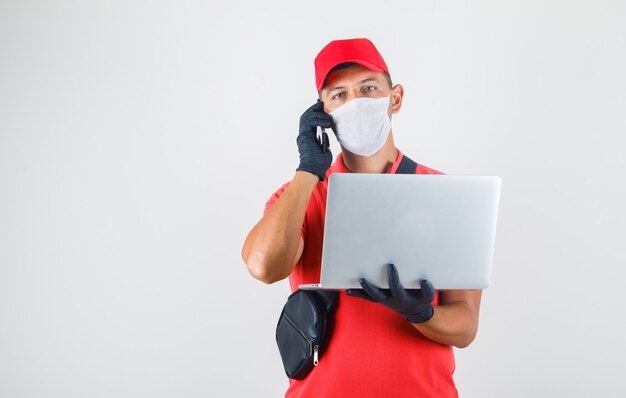 Delivery man holding laptop et parler au téléphone portable en uniforme rouge