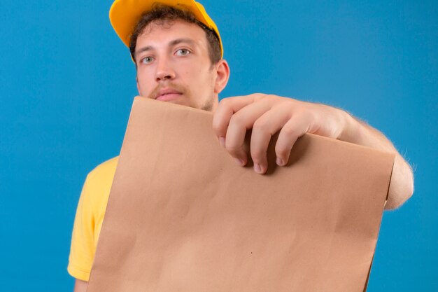 Delivery man in yellow polo et cap holding paper package looking at camera avec visage sérieux debout sur bleu isolé