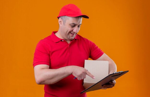 Delivery man wearing red uniform et cap holding box package et presse-papiers pointant avec l'index vers lui souriant sur mur orange isolé