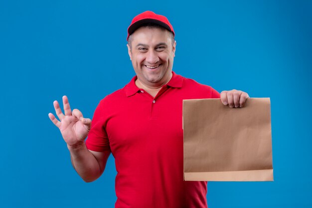 Delivery man wearing red uniform et cap holding paper package à positif et heureux de faire signe ok sur mur isolé bleu