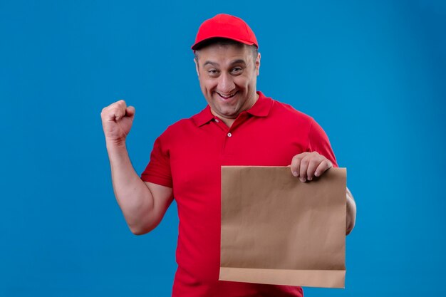 Delivery man wearing red uniform et cap holding paper package smiling face lever le poing après une victoire debout sur un espace bleu isolé
