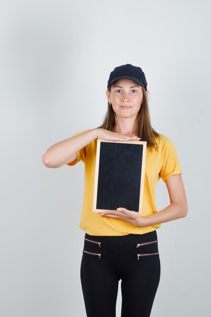 Delivery woman holding blackboard en t-shirt, pantalon et casquette et à la joie