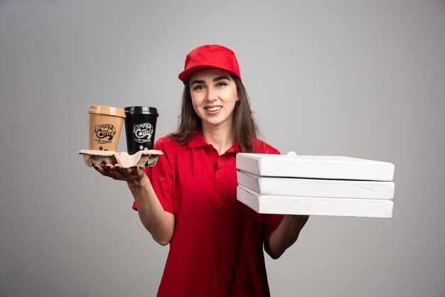 Delivery woman holding pizza et tasses à café sur mur gris.