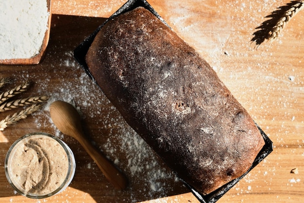 Démarreur actif de seigle et de levain de blé dans un bocal en verre à côté de l'ingrédient farine et pain de grains entiers fraîchement cuit Disposé sur la table