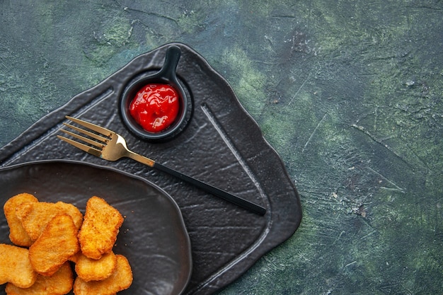 Photo gratuite demi-coup de pépites de poulet sur une plaque noire et de ketchup à la fourchette sur un plateau de couleur foncée