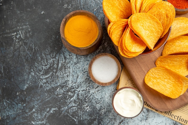 Demi-coup de savoureuses chips maison coupées en tranches de pommes de terre sur une planche à découper en bois et différentes épices sur du papier journal sur table grise