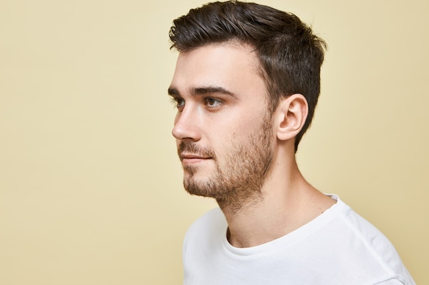 Demi-image de profil de beau jeune homme de race blanche avec une bonne peau, yeux bruns, cheveux noirs élégants et chaume posant isolé contre un mur blanc, regardant en face de lui, souriant