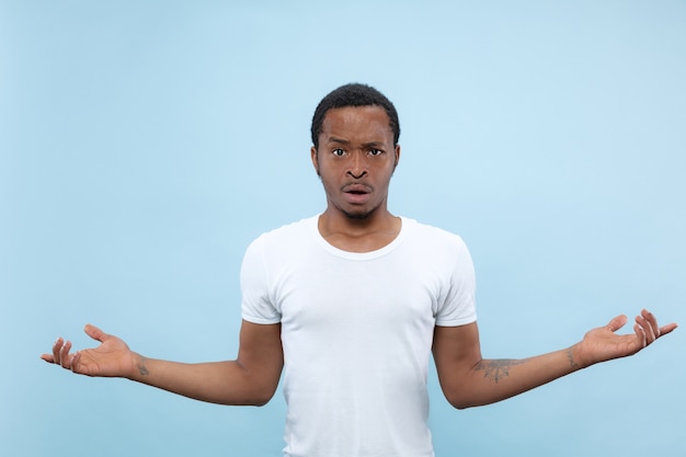 Demi-longueur gros plan portrait de jeune homme afro-américain en chemise blanche sur fond bleu. Émotions humaines, expression faciale, concept publicitaire. Demander et incertain, doutes, émotions négatives.