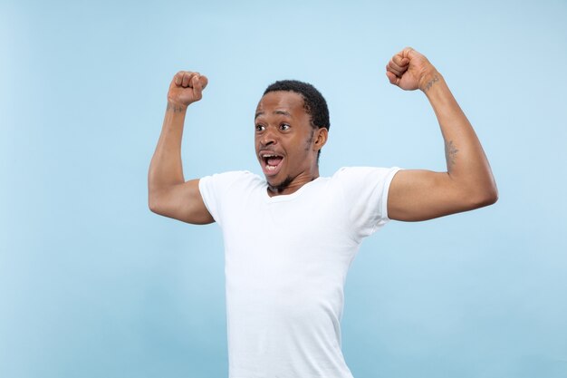 Demi-longueur gros plan portrait de jeune homme afro-américain en chemise blanche sur fond bleu. Émotions humaines, expression faciale, publicité, concept. Célébrer, émerveillé, étonné, choqué, fou heureux.