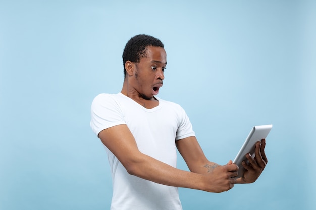 Demi-longueur gros plan portrait de jeune homme afro-américain en chemise blanche sur mur bleu. Émotions humaines, expression faciale, publicité, ventes, concept. Utilisation de la tablette, choqué, étonné.