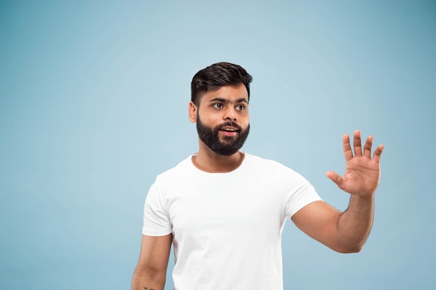 Demi-longueur gros plan portrait de jeune homme hindou en chemise blanche sur fond bleu. Émotions humaines, expression faciale, concept publicitaire. Espace négatif. Affichage de la barre d'espace vide, pointage, salutation.