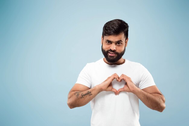 Demi-longueur gros plan portrait de jeune homme hindou en chemise blanche sur fond bleu. Émotions humaines, expression faciale, concept publicitaire. Espace négatif. Montrant le signe du cœur. Souriant.