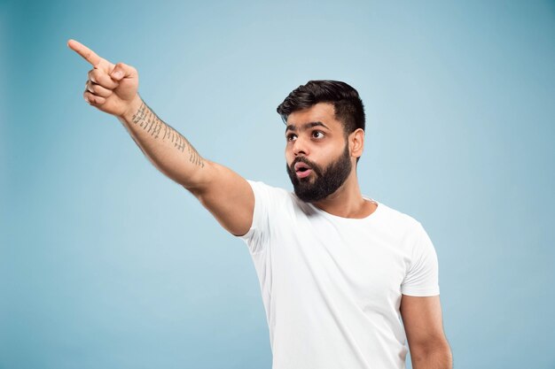 Demi-longueur gros plan portrait de jeune homme hindou en chemise blanche sur fond bleu. Émotions humaines, expression faciale, ventes, concept publicitaire. Espace négatif. Pointant du doigt être choqué et étonné.