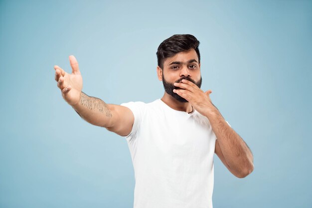 Demi-longueur gros plan portrait de jeune homme hindou en chemise blanche sur fond bleu. Émotions humaines, expression faciale, ventes, concept publicitaire. Espace négatif. Pointant vers le haut, être heureux et étonné.