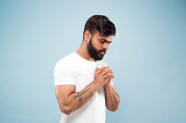 Demi-longueur Gros Plan Portrait De Jeune Homme Hindou En Chemise Blanche Isolé Sur Fond Bleu. émotions Humaines, Expression Faciale, Concept Publicitaire. Espace Négatif. Debout Et Priant Les Yeux Fermés.