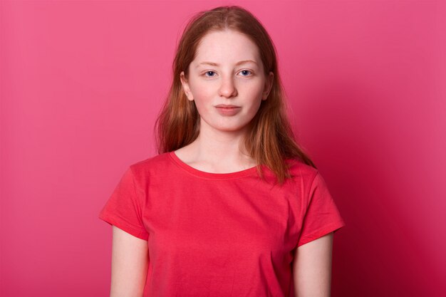 Demi-longueur jeune étudiante regarde avec une expression faciale calme, portant un t-shirt décontracté rouge, a de longs cheveux bruns raides et des yeux bleus, isolé sur rose