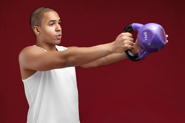 Demi-portrait de profil de beau jeune bodybuilder à la peau sombre avec tête rasée exerçant avec des haltères. Sportif africain musclé travaillant avec un poids lourd, renforçant les muscles des bras