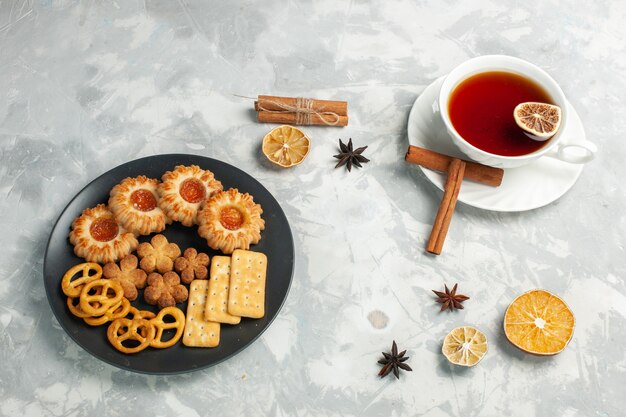 Demi-vue de dessus de délicieux cookies avec des craquelins et des chips à l'intérieur de la plaque avec une tasse de thé sur le bureau blanc clair