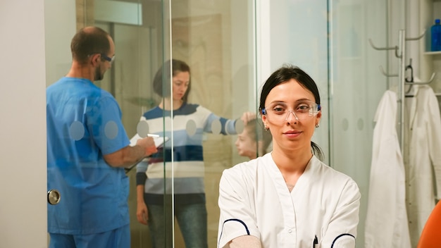 Dentiste pédiatrique regardant la caméra en souriant, tandis que l'homme assiste et parle avec les patients de l'hygiène dentaire en arrière-plan. Stomatologue assis devant une webcam travaillant dans une clinique dentaire