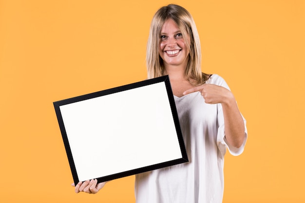 Photo gratuite désactiver le doigt pointé jeune femme blonde au cadre photo blanc vide