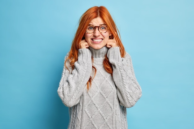 Désactivez le son. La jeune femme aux cheveux roux mécontente serre les dents, bouche les oreilles pour éviter le bruit ennuyeux ne veut pas écouter de la musique très forte porte un chandail d'hiver chaud.