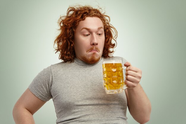 Désemparé jeune homme aux cheveux roux aux cheveux bouclés tenant un verre de bière légère, le regardant, ayant confus l'expression indécise, hésitant, pensant boire ou non, debout contre un mur blanc