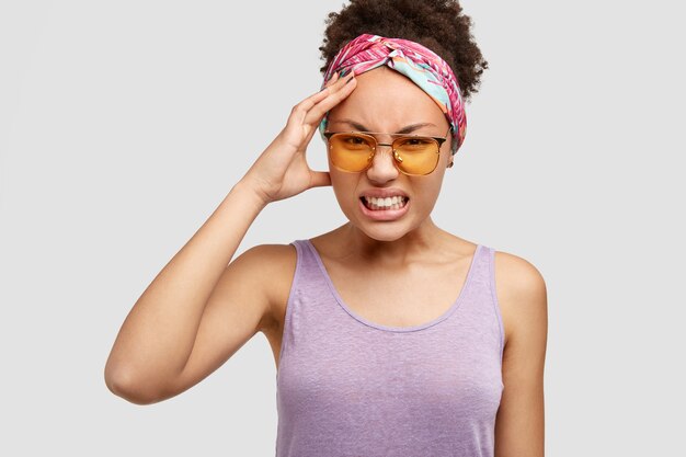 Désespérée, jeune femme afro-américaine fronce les sourcils face à l'insatisfaction, serre les dents, garde la main sur la tête, a mal à la tête, pose contre le mur blanc. Femme séduisante à la peau foncée irritée