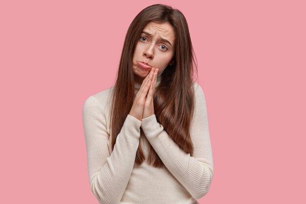 Désespérée et triste jeune femme charmante aux cheveux longs, garde les paumes dans un geste de prière, demande à Dieu de réaliser ses rêves, demande grâce