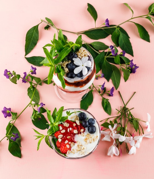 Dessert avec des fraises, des bleuets, des noix, de la menthe, des branches de fleurs dans un gobelet et un vase sur une surface rose, à plat.