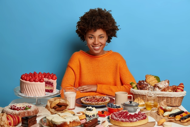 Desserts, restauration rapide, concept de mode de vie malsain. Heureux modèle à la peau sombre en pull orange, aime faire la fête, n'a pas de régime, stimule l'humeur avec des plats sucrés, isolés sur un mur bleu.