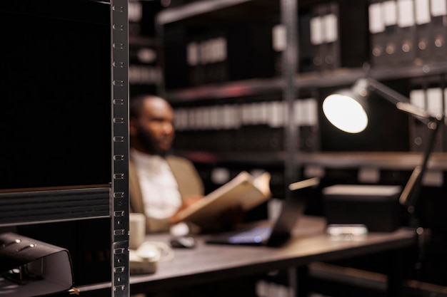 Photo gratuite détective afro-américain travaillant dans une pièce sombre éclairée par une lampe de bureau. flic flou assis à la table du lieu de travail et analysant les preuves du dossier de crime la nuit mise au point sélective