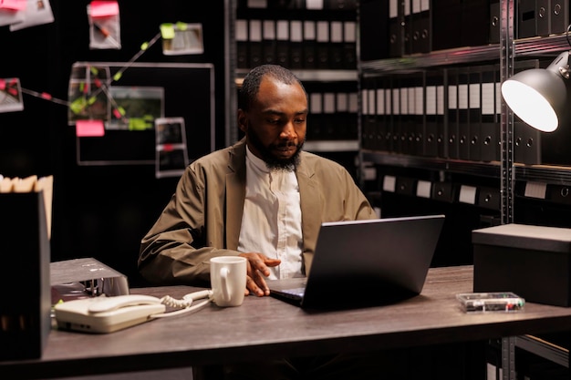 Photo gratuite détective privé assis à son bureau et travaillant sur son ordinateur portable dans l'agence la nuit. criminaliste afro-américain concentré à la recherche de dossiers de crimes sur ordinateur dans le bureau du détective de la police