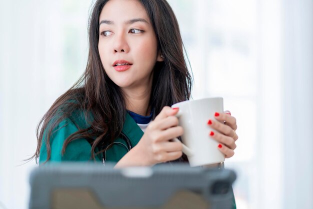 Détendez-vous après le travail femme médecin asiatique main tenir une tasse de café chaud décontracté se détendre après avoir travaillé dur la télémédecine en clinique tout en portant l'uniforme