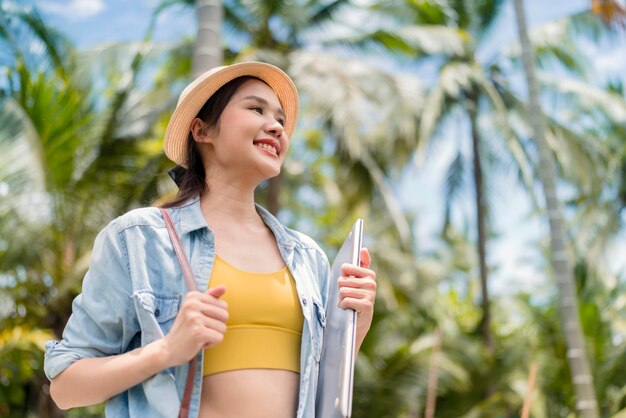 Détendez-vous décontracté insouciant souriant asiatique femme nomade numérique programmeur indépendant main tenir un ordinateur portable marchant à la plage et cocotier bleu ciel nouveau style de vie millénaire travaillant n'importe où avec bonheur