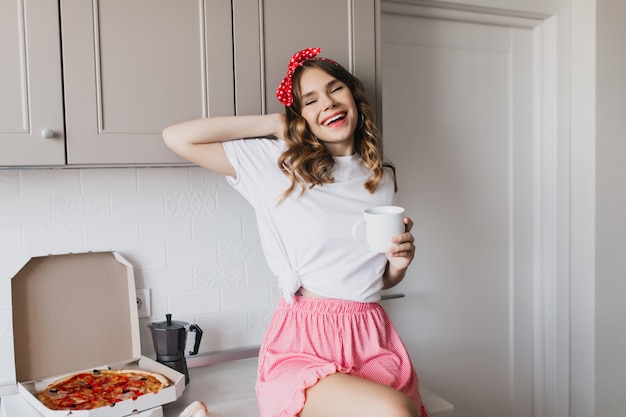 Détendu jeune femme passant la matinée avec une tasse de café et de pizza. Fille bouclée raffinée jouant avec ses cheveux tout en posant dans la cuisine.