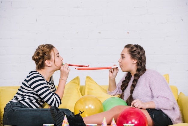 Deux adolescentes soufflant corne de parti assis face à face sur le canapé avec des ballons