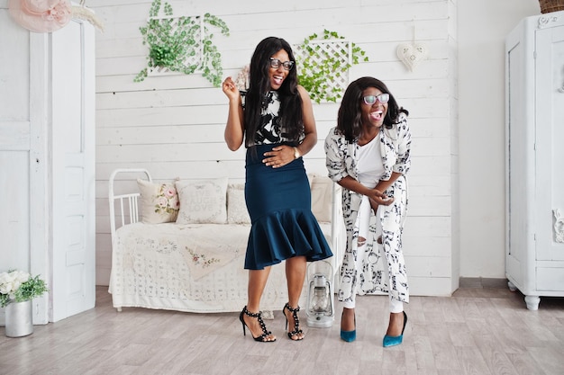 Photo gratuite deux amies africaines portent des lunettes posées à l'intérieur de la salle blanche rire et s'amuser ensemble