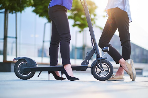 Deux amis discutent dans la rue par une belle journée ensoleillée. L'une d'elles roule en scooter électrique et porte des talons.