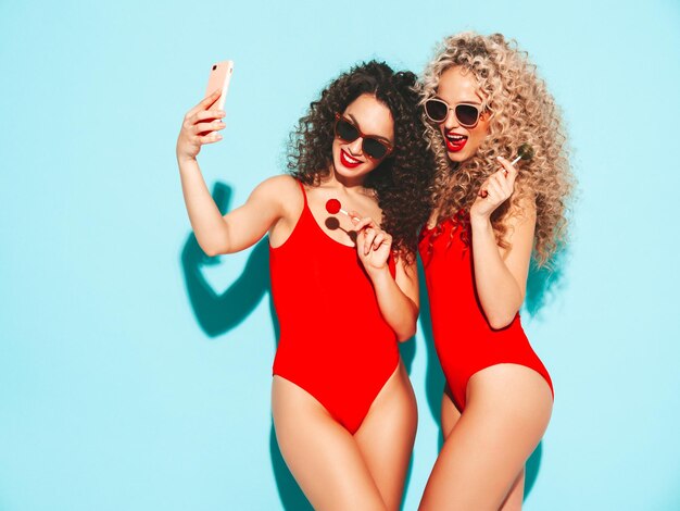Deux belles femmes hipster souriantes en maillot de bain rouge Modèles à la mode avec une coiffure bouclée prenant un selfie en studio Femme chaude posant près d'un mur bleu Tenant une sucette Prendre des photos de selfie
