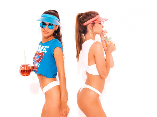 Deux belles femmes sexy souriantes en sous-vêtements d'été blancs et sujet. Filles à la mode en lunettes de soleil, casquette visière transparente. Devenir fou. Modèles drôles isolés. Boire une boisson fraîche de cocktail