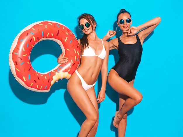 Deux belles femmes souriantes sexy en maillot de bain d'été blanc et noir maillots de bain.Filles à lunettes de soleil. Modèles positifs s'amusant avec un matelas gonflable donut lilo isolé sur mur bleu