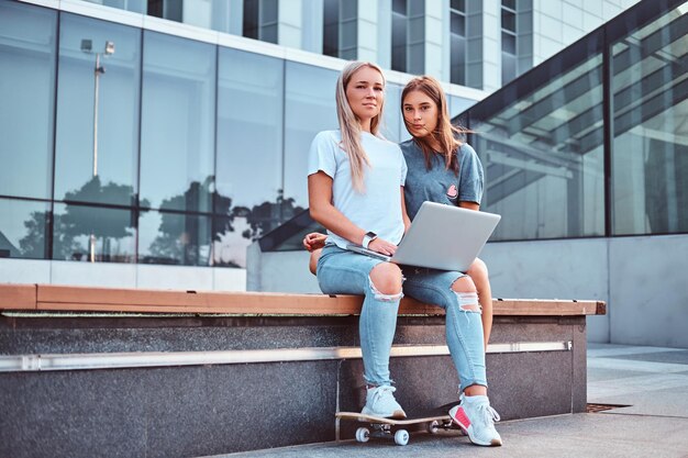 Deux belles filles hipster assises sur le banc avec un ordinateur portable sur fond de gratte-ciel.