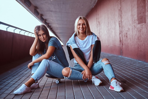 Deux belles filles hipster heureuses assises sur une planche à roulettes sur un trottoir sous le pont.