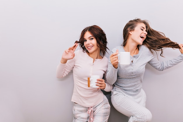 Deux Belles Filles En Pyjama S'amusant Sur Un Mur Gris. Fille Aux Cheveux Longs En Riant Et Garde Les Yeux Fermés, L'autre Aux Cheveux Bouclés En Souriant.